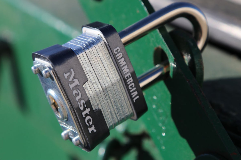 A Master Lock No. 3 laminated steel padlock locked through the hasp of a green dumpster, beaded with water.